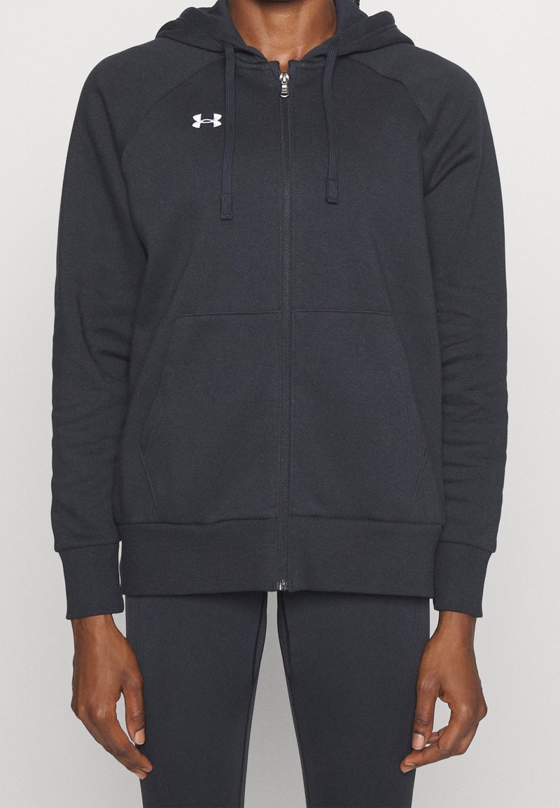Personne portant un sweat à capuche zippé noir avec un petit logo blanc Under Armour sur la poitrine et un pantalon noir assorti sur un fond uni.