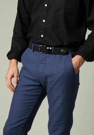 Man draagt een zwart, geknoopt overhemd, ingestopt in een blauwe broek met een zwarte gevlochten riem, één hand in de broekzak, de andere ontspannen langs zijn zijde.