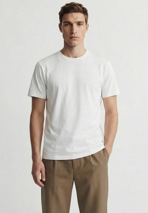 Jeune homme portant un t-shirt blanc uni à manches courtes et un pantalon marron, debout devant un fond clair neutre.