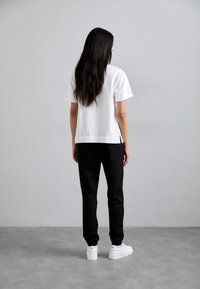 Hvit bomulls T-skjorte med korte ermer, kombinert med svarte joggebukser. Modellen har på seg hvite sneakers, sett fra baksiden mot en grå bakgrunn.