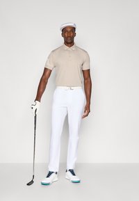 Beige polo skjorte med teksturerte skulderdeler, hvite bukser og hvite sko med marineblå detaljer, sammen med en hvit caps og golfkølle.