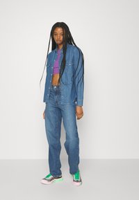 Jeansjacke und Jeans in Mittelblau, eng geschnitten mit zwei Brusttaschen; kombiniert mit einem lila Crop-Top und mehrfarbigen Sneakern.