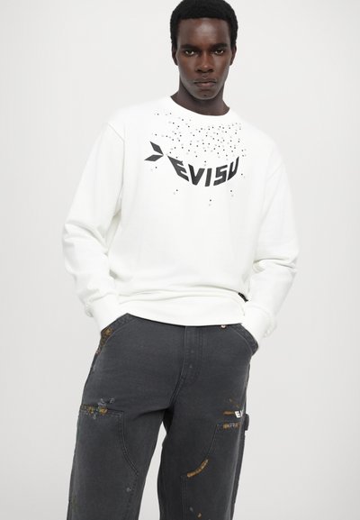Hvid sweatshirt med sort grafisk tekst og prikker, parret med mørkegrå cargo-bukser med slidte detaljer og farverige accenter.