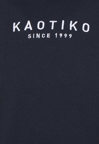 Tmavě modré bavlněné tričko s bílým vyšívaným nápisem "KAOTIKO" a "SINCE 1999" umístěným na středu hrudi. Hladká textura, ležérní design.