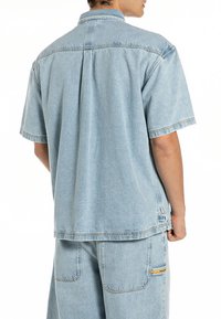 Camisa de ganga azul clara de manga curta com um ajuste descontraído, colarinho abotoado e costura na pala das costas. Apresenta costuras subtis e sem padrões visíveis.