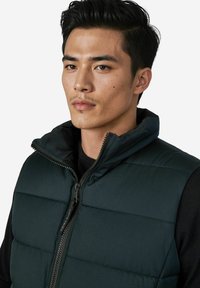 Dunkelgrüne, wattierte Weste mit hohem Kragen, ausgestattet mit einem Frontreißverschluss und glatter Textur. Wird über einem schwarzen Hemd getragen.