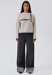 Maglione in maglia crema con motivi a treccia e dettagli neri, abbinato a jeans scuri a gamba larga con una finitura grigia.