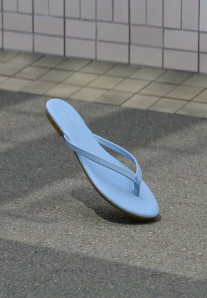 Lichtblauwe teenslipper die boven het asfalt zweeft nabij een betegelde muur en een stoep.