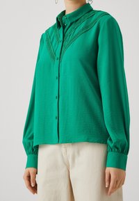 Blusa verde con bottoni realizzata in tessuto testurizzato, con cuciture decorative lungo le spalle e maniche lunghe a palloncino.