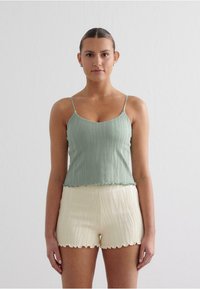 Camisetă verde deschis, cu nervuri, cu bretele subțiri și margine ondulată, asortată cu pantaloni scurți de culoare crem, cu textură și margine sinuoasă.
