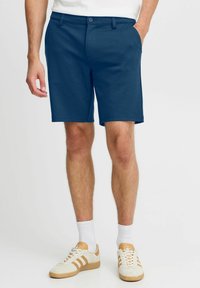Marineblå shorts laget av lett stoff, med knappelukking, sidelommer og skreddersydd passform, kombinert med hvite og brune joggesko.