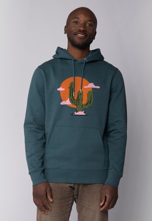 Man met een tealkleurige hoodie met een afbeelding van een cactus en een oranje zon, staand tegen een effen lichtgrijze achtergrond.