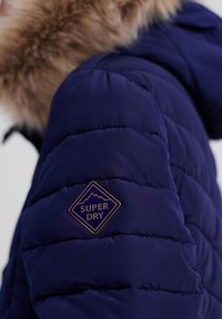 Marineblå puffjakke med en faux fur hætte, der har et diamantformet "Superdry" logo patch på ærmet og kviltet tekstur.