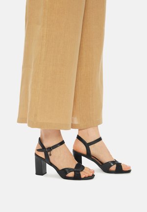 Černé kožené sandály na vysokém podpatku s otevřenou špičkou, vybavené překříženými pásky a přezkou u kotníku, v kombinaci s hnědými kalhotami s širokými nohavicemi.