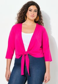 Cardigan fuchsia à nouer devant avec des manches trois-quarts, porté sur un top blanc, associé à un jean en denim foncé. Tissu lisse, design décontracté.