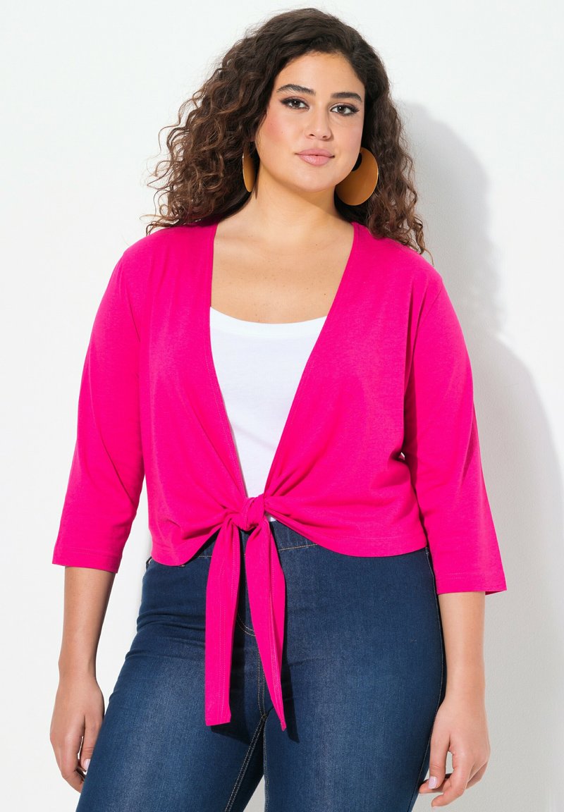 Cardigan fuchsia à nouer devant avec des manches trois-quarts, porté sur un top blanc, associé à un jean en denim foncé. Tissu lisse, design décontracté.
