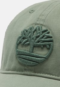 Gorra de algodón verde con un logo de árbol bordado texturizado. Presenta un visera curva y detalles cosidos en la tela.