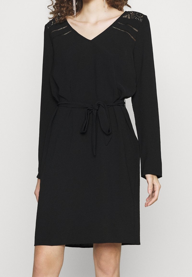 Femme portant une robe noire mi-longue avec manches longues, col en V, détail en dentelle aux épaules, et une ceinture en tissu nouée à la taille.