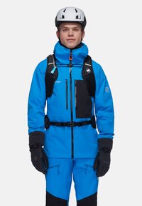Giacca da sci blu brillante con accenti neri, dotata di un colletto alto, multiple tasche con zip, polsini regolabili e guanti; indossata sopra pantaloni coordinati.