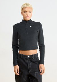 Czarny krótki top z długim rękawem i zamkiem przy kołnierzu, wykonany z teksturowanej tkaniny, z małym białym logo Nike na lewej piersi.