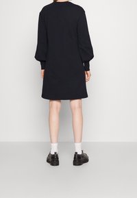 Abito felpa oversize nero con maniche lunghe a palloncino e polsini a coste. Orlo piatto, design semplice, indossato con scarpe nere e calze bianche.