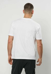 Mann med kort hår iført en enkel hvit t-skjorte og svarte bukser, stående med ryggen mot kameraet foran en ensfarget lys bakgrunn.