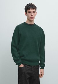 Dunkelgrüner, gerippter Pullover mit Rundhalsausschnitt, lässiger Passform und langen Ärmeln, kombiniert mit dunklen Jeans. Glatte Textur und legeres Design.