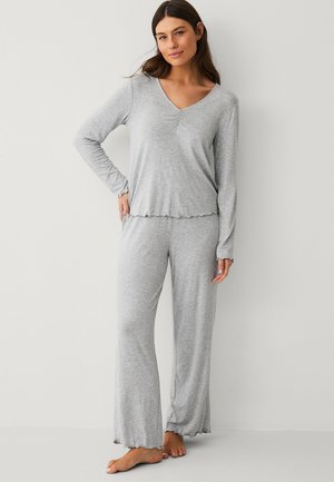 Kvinde stående barfodet iført lysegrå langærmet top og løstsiddende pyjamasbukser, poserer med den ene hånd på hoften mod en ensfarvet baggrund.