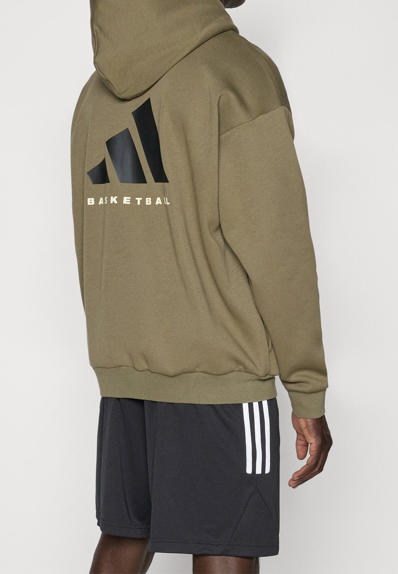 Personne portant un sweat à capuche vert olive avec le mot « BASKETBALL » et un logo au dos, associé à un short noir avec trois bandes blanches.