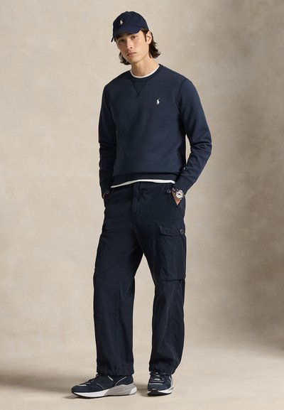 Giovane uomo con felpa blu navy, pantaloni cargo, cappellino e scarpe da ginnastica, in piedi con le mani nelle tasche davanti a uno sfondo neutro.