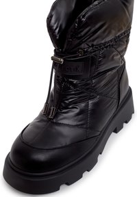 Botín negro acolchado hasta el tobillo con un acabado brillante, que cuenta con una suela de goma, textura acolchada, cordones ajustables y una etiqueta de logo en el lateral.