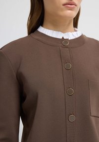 Brown cotton blend garment with a round neckline, featuring a button-up front, four metallic buttons, and a pocket. Textured fabric with a ribbed collar.