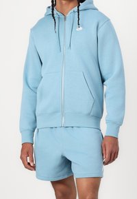 Felpa con zip azzurra chiara realizzata in tessuto morbido, con una tasca frontale e un piccolo logo Nike bianco sul petto. Abbinata a pantaloni sportivi coordinati.