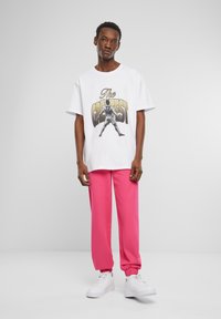 Hvid grafisk t-shirt med teksten "The Greatest" og en illustration, parret med lyserøde sweatpants og hvide sneakers, stående foran en neutral baggrund.