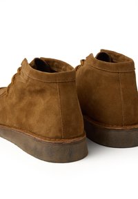 Bruine suède enkelboots met ronde hakken, bruine veters en een gestructureerde rubberen zool. Beschikt over een gladde afwerking met subtiele stikdetails.