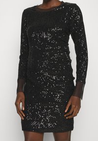 Svart paljettklänning med långa ärmar, strukturerad tyg och åtsittande form; har en genomskinlig mesh-krage och en upphöjd nederkant.