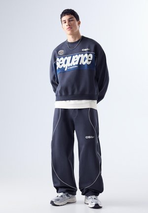 Tânăr bărbat poartă un hanorac larg, de culoare închisă, cu textul "Sequence", pantaloni largi asortați, adidași albi și un lanț argintiu la gât.