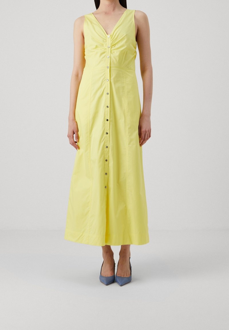 Žluté, bezrukávové maxi šaty s výstřihem do V, nařasením v oblasti dekoltu a předním zapínáním na knoflíky. Nosit s modrými podpatky.