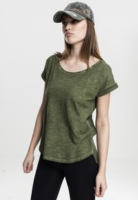 T-shirt a maniche corte verde oliva con risvolti, realizzato in un tessuto testurizzato. Indossato con leggings neri e un cappellino camouflage.