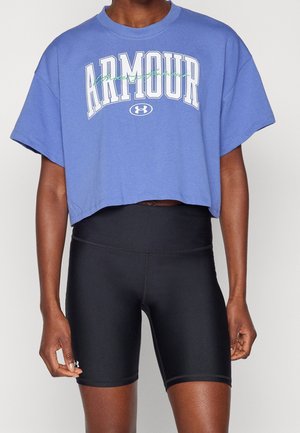 Person iført en blå cropped Under Armour t-shirt og sorte tætsiddende høj-taljerede træningsshorts mod en ensfarvet baggrund.