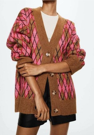 Brun cardigan med pink og sort argyle mønster, store knapper, båret over hvid top og sorte shorts, arme krydset foran.