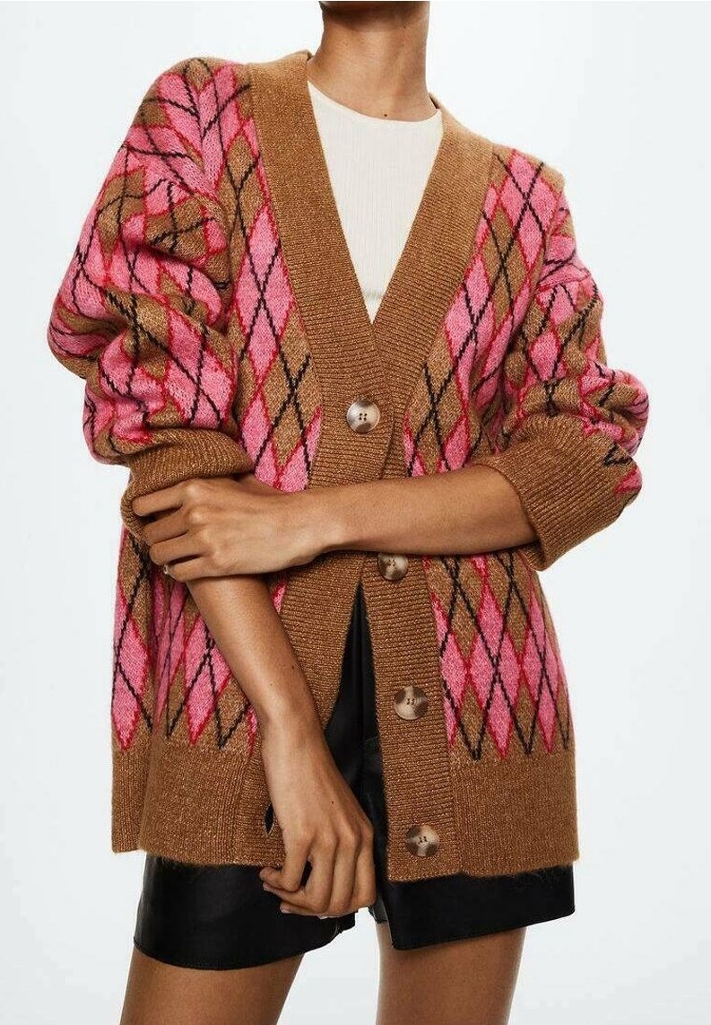 Cardigan marron avec un motif argyle rose et noir, gros boutons, porté sur un haut blanc et un short noir, bras croisés devant.