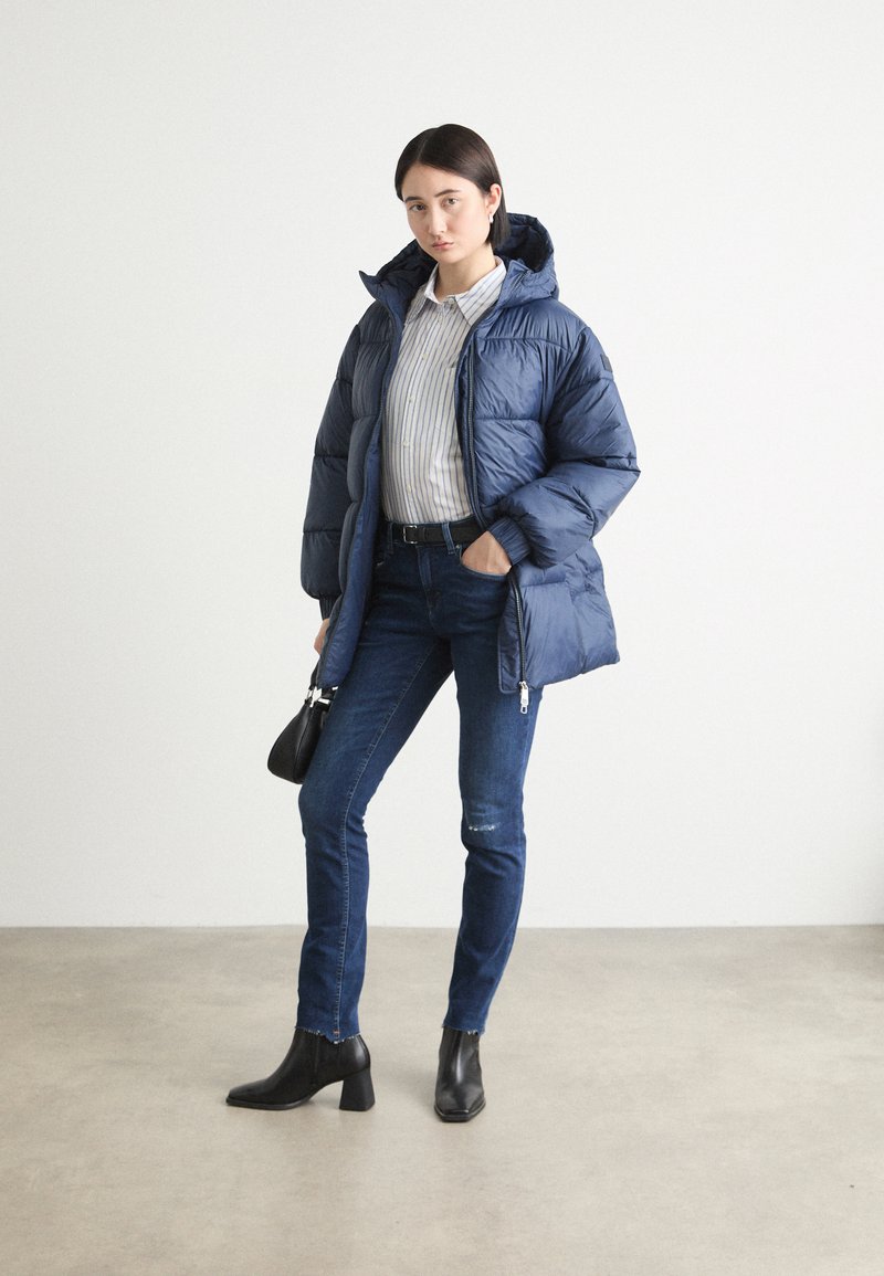 Chaqueta de plumón azul marino con capucha, superficie texturizada, combinada con una camisa de rayas clara, jeans oscuros y botas negras de tobillo. Incluye un bolso pequeño.
