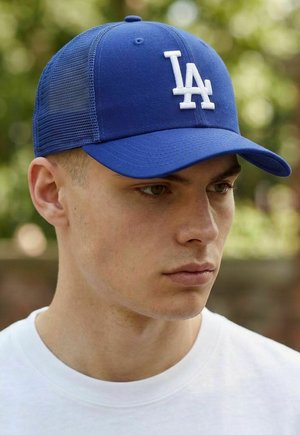 Junger Mann mit blauem Baseballcap und weißem "LA"-Logo sowie weißem Hemd, im Freien vor unscharfem natürlichem Hintergrund.