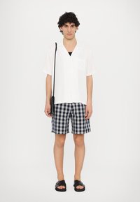 Hvit kortermet skjorte, marineblå og hvite rutete shorts, svarte sandaler og en svart crossover-bag. Enkle, avslappede strandklær.