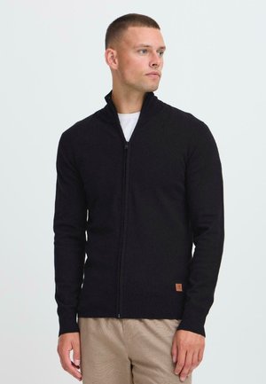 Hombre vestido con un cárdigan negro con cremallera sobre una camisa blanca y pantalones beige, mirando hacia la derecha contra un fondo claro y liso.