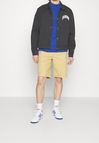 Czarna kurtka z przednimi kieszeniami, niebieska koszulka, khaki szorty i białe sneakersy z niebieskimi i czerwonymi akcentami. Model stoi na neutralnym tle.