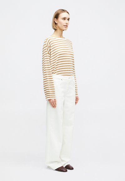 Femme aux cheveux blonds courts portant un haut à manches longues à rayures marron et blanc, un pantalon ample blanc et des chaussures marron foncé debout.