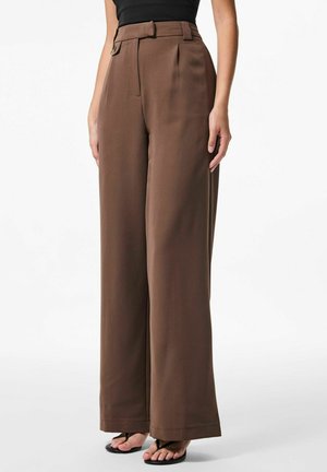 Bruine wijde broek met hoge taille, voorzien van riemlussen, plooiingen en een gladde textuur. Gecombineerd met zwarte flip-flops.
