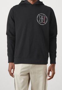 Sudadera de algodón negra con un bolsillo frontal, capucha con cordón y un logo que presenta una prominente letra "H" en blanco y rojo. Textura suave.